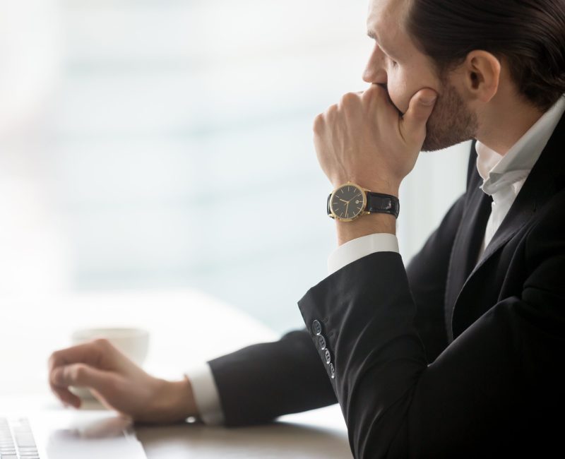 Serious concerned young businessman looks towards the window in deep thought sitting at workplace in modern office. Thinking about corporate future, work problem resolution, tough decision concept.