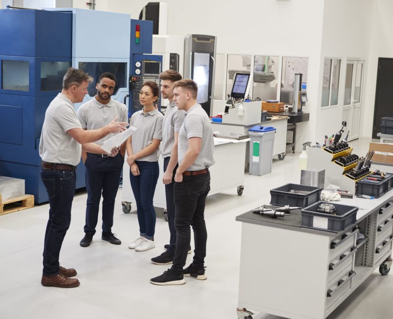 Engineering Team Meeting On Factory Floor Of Busy Workshop